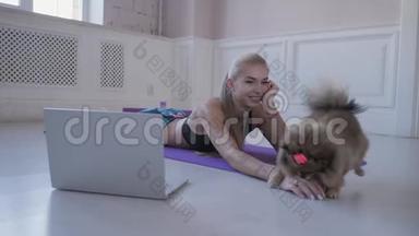 家庭健身。 年轻女子在运动和玩小狗斯皮兹。 在网上观看笔记本电脑上的视频